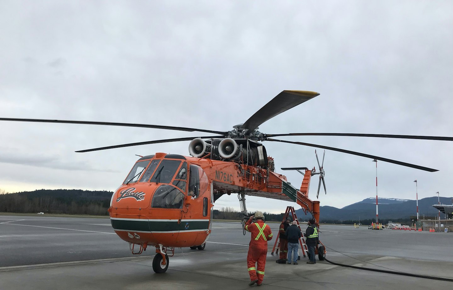 Aviation technicians refuel a large orange helicopter on an airport tarmac.