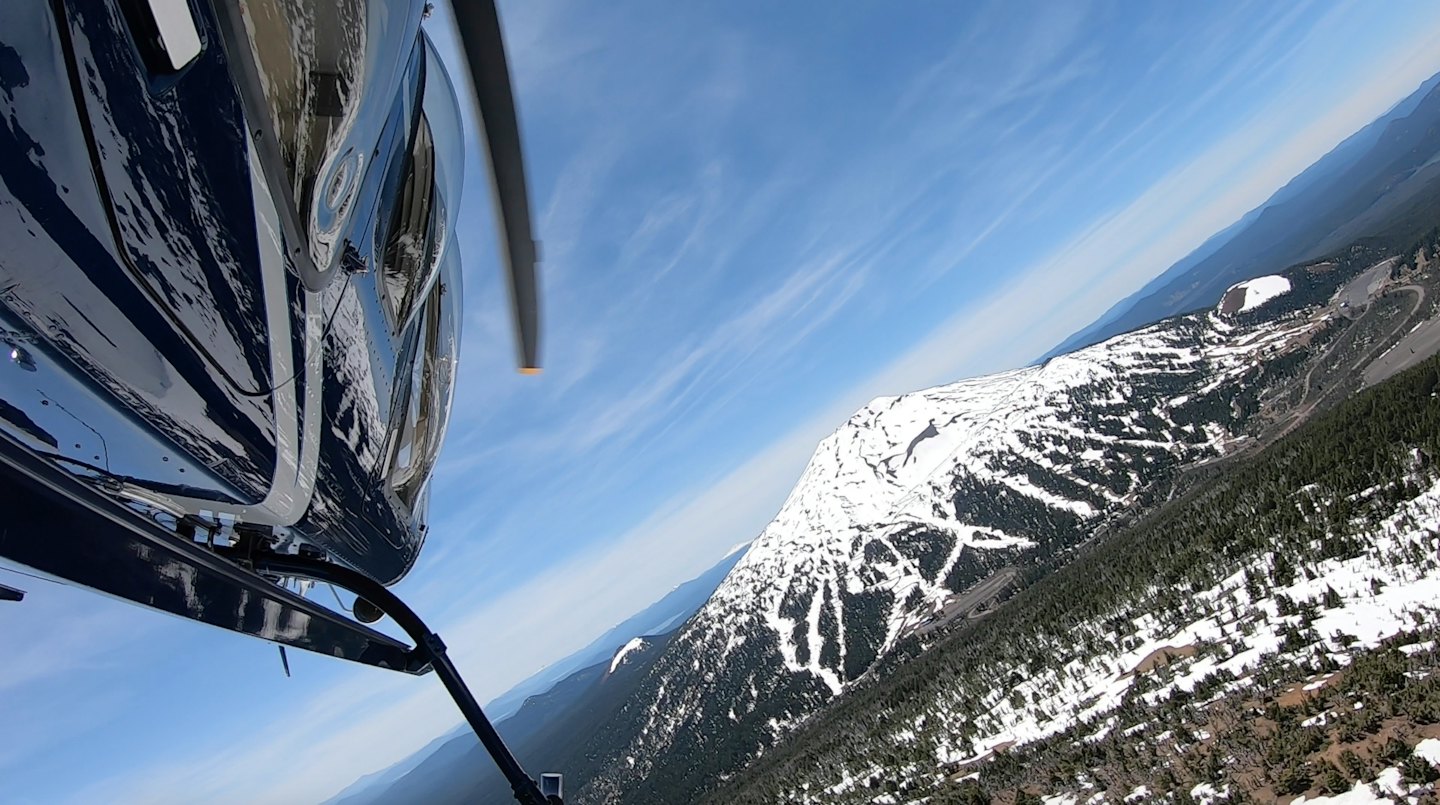 Helicopter flying over snow-covered mountains and forests.